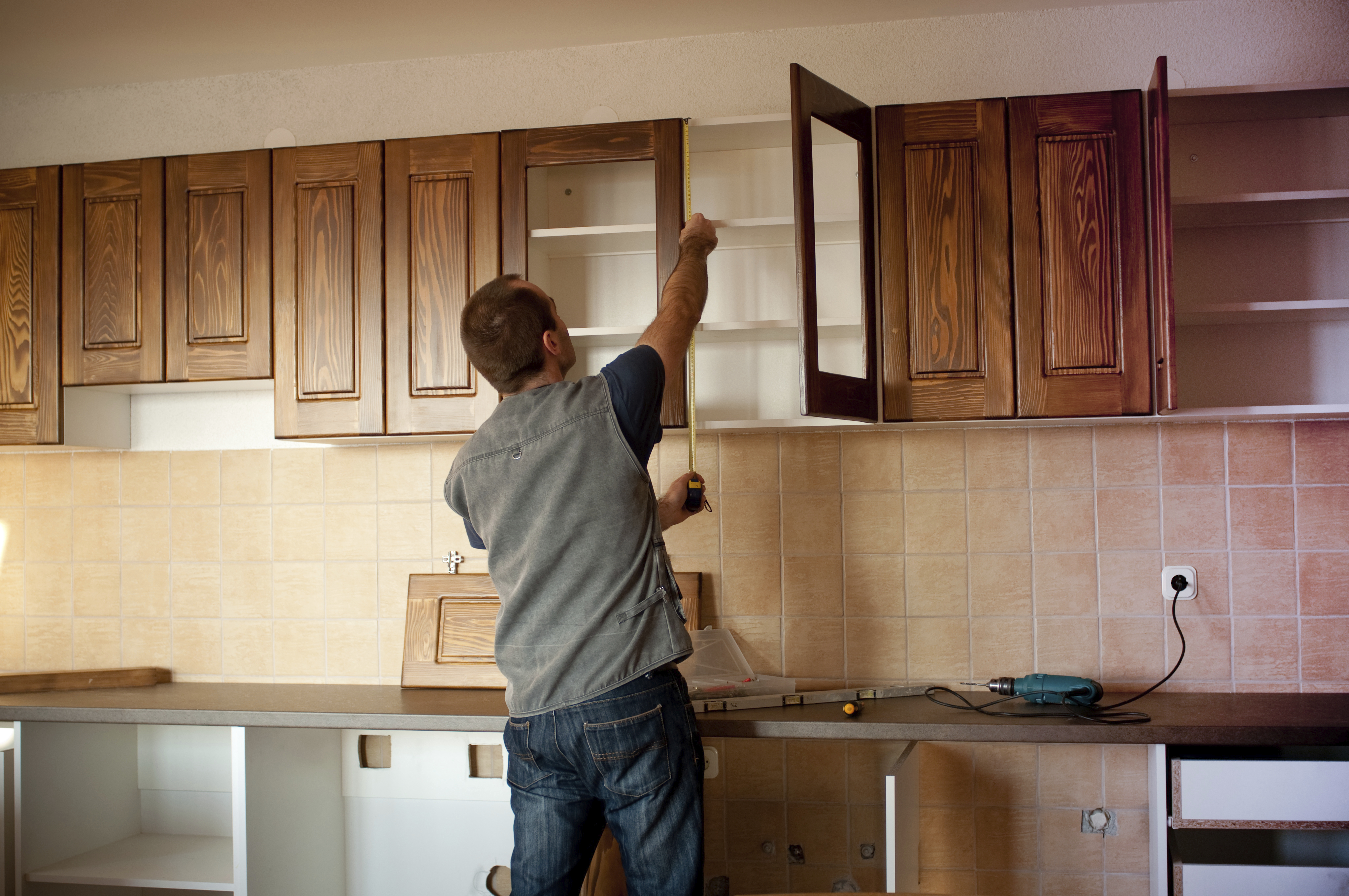 A man in a kitchen measuring brown wooden kitchen cupboards.