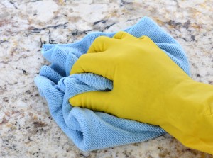 Closeup of a hand in a yellow latex glove using a blue towel to clean a granite counter top.